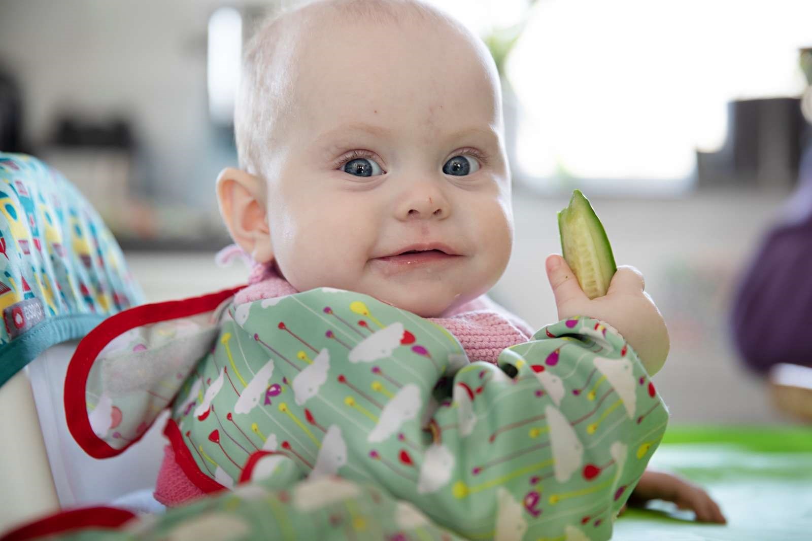 Billede af en baby i en spisesituation med et stykke agurk i hånden.